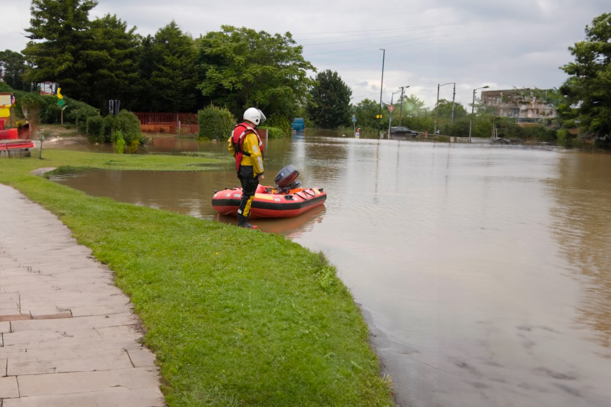 Paysage urbain inondé avec un bateau gonflable des services d'urgence en première ligne. Un secouriste en équipement se tient debout à côté du bateau, prêt à intervenir dans les zones touchées par les crues.